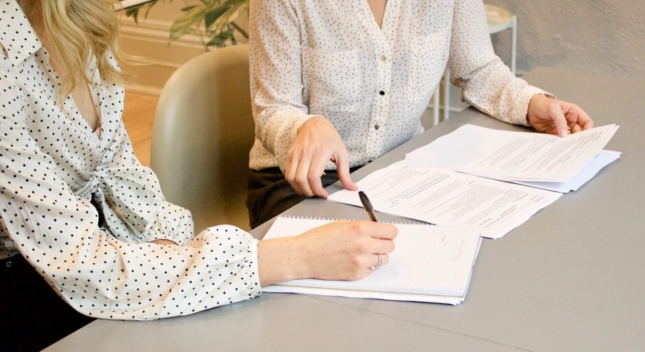 woman signing on white printer paper beside woman about to touch the documents